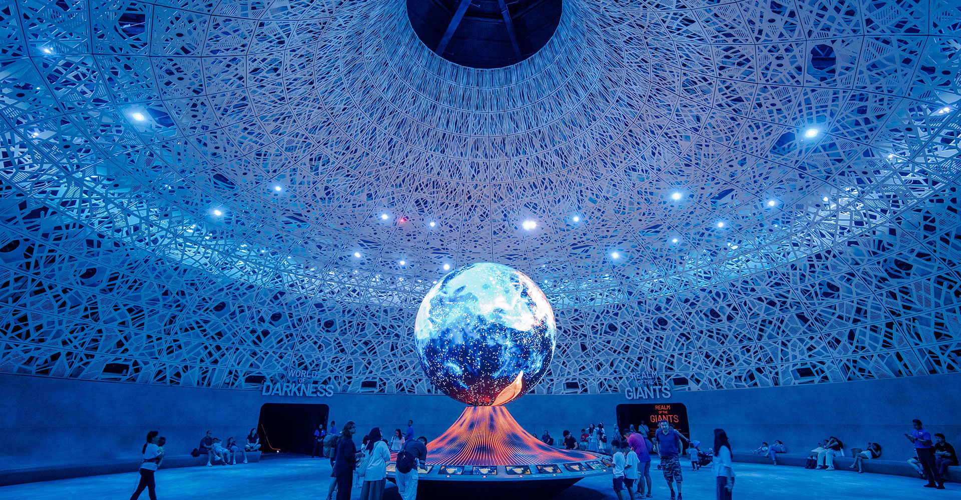 Large room with a globe in its center around which are gathered a dozen people, the ceiling curving inwards at the center.