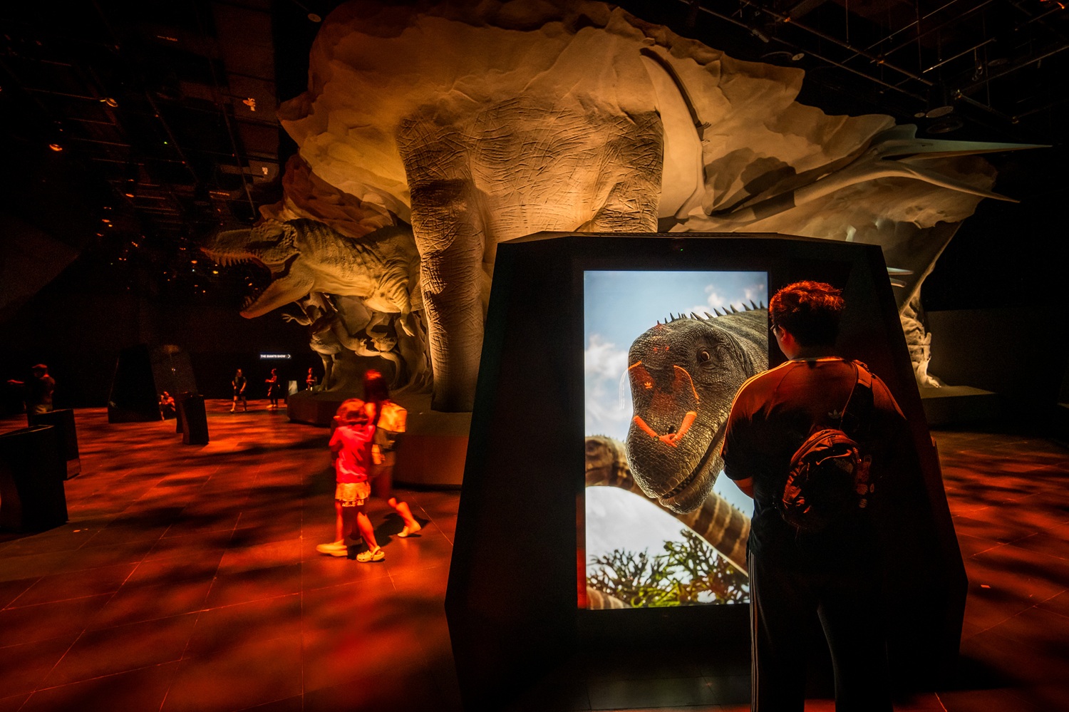 Dark room with statues of rock-embedded dinosaurs and a person standing in front of a large screen showing the face of a dinosaur.