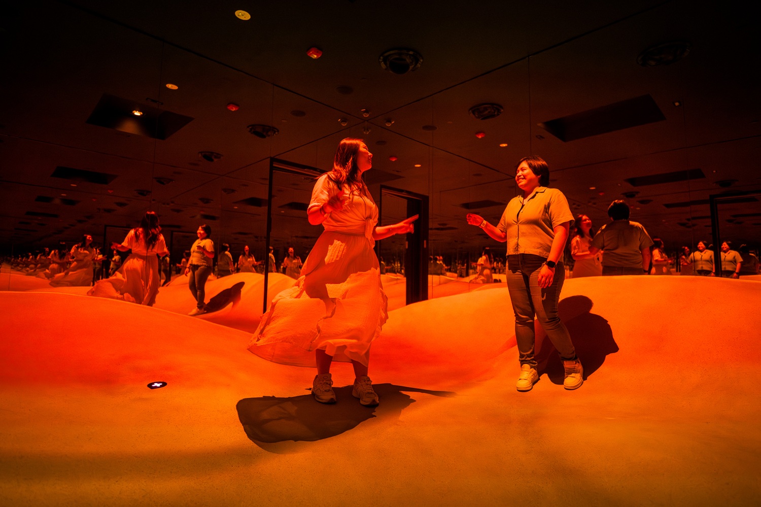Bright orange room with small dunes as the floor, each wall is a mirror, two people conversing and smiling in it.